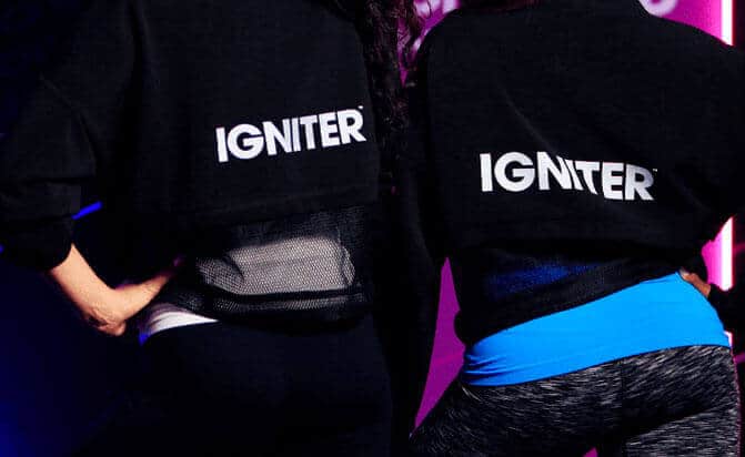 Two women in athletic clothing wear black sweatshirts with "IGNITER" on the back in white text